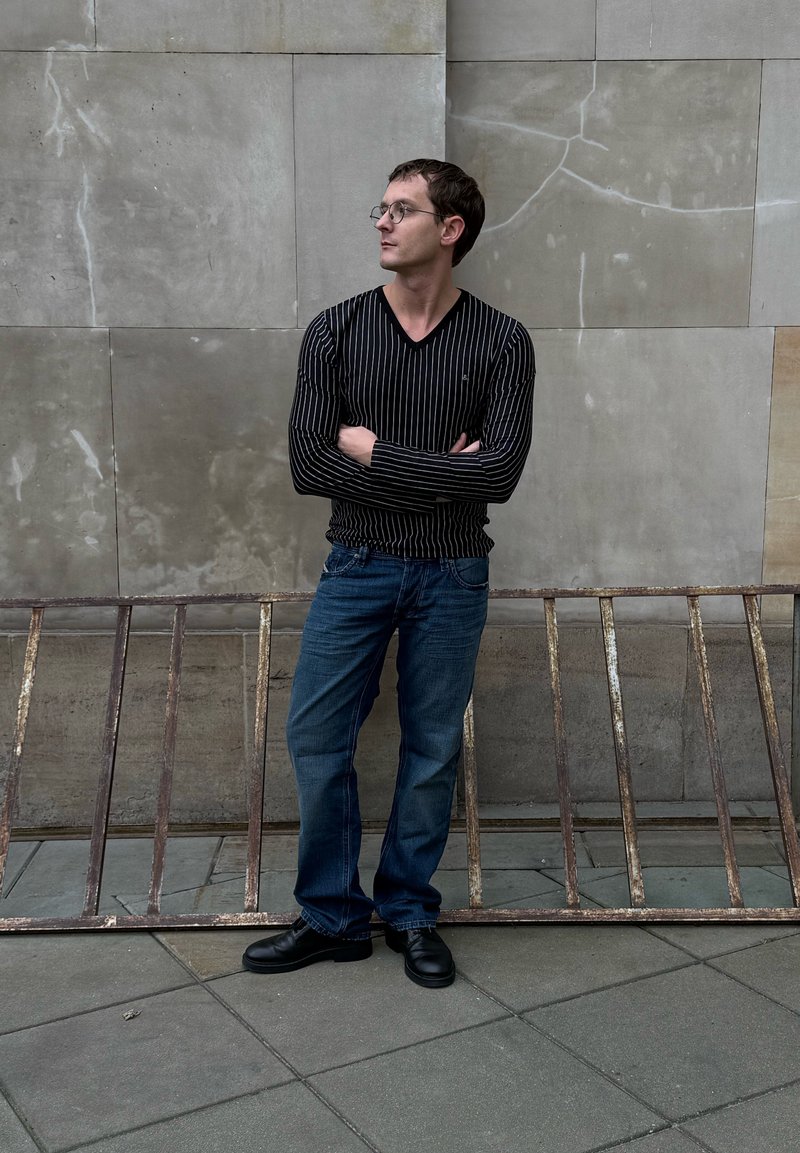 Chemise à manches longues rayée noire, jean bleu et chaussures noires. Debout contre un mur gris avec une rambarde métallique rouillée au premier plan.