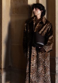 Jeune personne portant un long manteau à imprimé léopard et tenant une grande pochette brun foncé texturée, debout contre un mur en pierre.