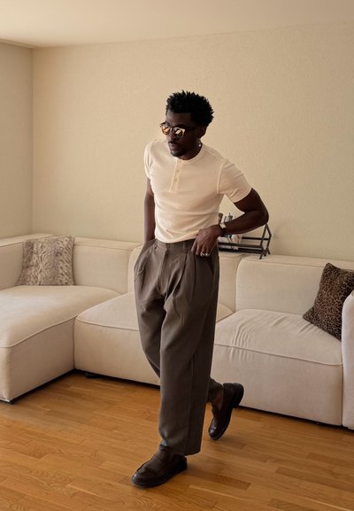 Hombre con gafas de sol, camisa crema de manga corta, pantalones plisados grises y zapatos negros, de pie en una sala de estar beige con un sofá seccional.