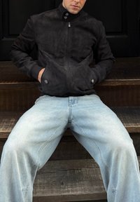 Fekete szarvasbőr cipzáras dzseki magas gallérral, két elülső zsebbel és ezüstszínű fém kiegészítőkkel, világoskék, laza szabású farmerrel párosítva.