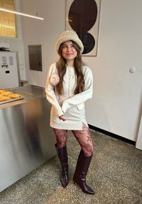 Jeune femme portant un chapeau crème, une robe-pull et des collants en dentelle avec des bottes marron, souriant dans une boulangerie près de plateaux de pain.