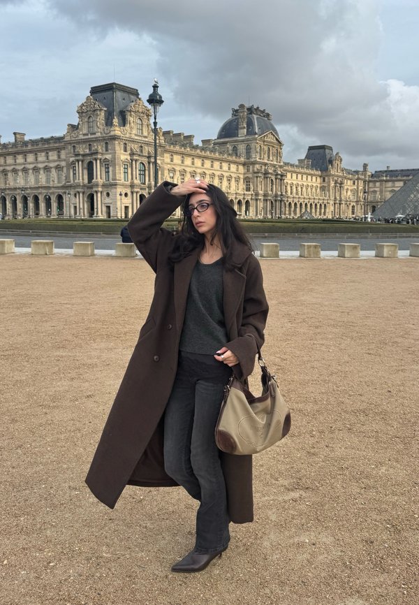 Femeie cu păr închis la culoare și ochelari, purtând o haină lungă maro și ținând o geantă bej, stă în fața Muzeului Louvre și a piramidei de sticlă, sub un cer înnorat.