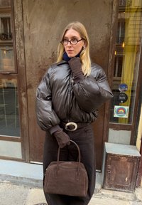 Schwarze, wattierte Jacke, dunkle Handschuhe und hoch taillierte schwarze Hose. Braune Handtasche, große runde Brille, mit einem blauen Schal um den Hals.