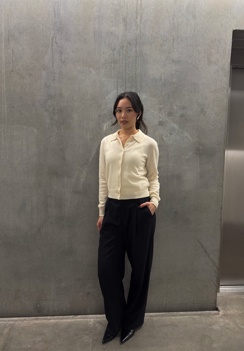 Donna in un cardigan crema chiaro, pantaloni neri a gamba larga e décolleté neri affusolati; si trova appoggiata a un muro grigio texturizzato.