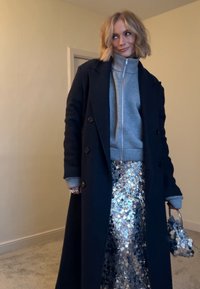 Manteau en laine bleu marine porté sur un sweat à capuche gris à fermeture éclair, associé à une jupe argentée à sequins. Porte un petit sac métallique. Détails texturés et brillants.