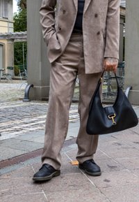 Μπεζ σουέτ κοστούμι με διπλό κουμπί και φαρδιές παντελόνες, σε συνδυασμό με μια μαύρη δερμάτινη τσάντα χειρό�ς με χρυσά αξεσουάρ και λεία υφή.