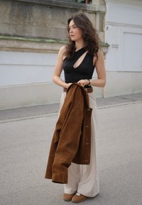 Zwarte mouwloze top met een uitsnijding, beige wijde broek, en een bruine suède jas in de hand. Bruine sneakers met witte accenten.