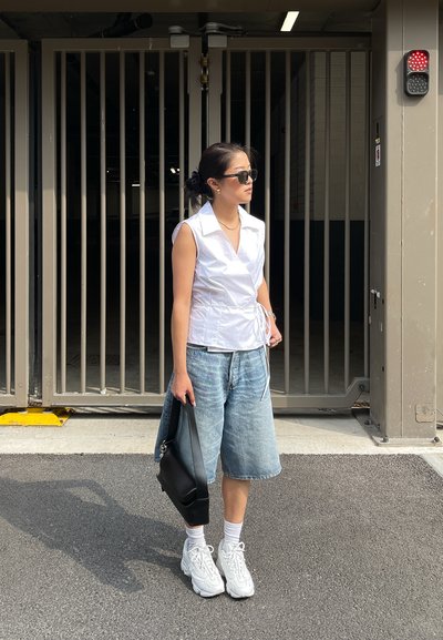 Top blanco sin mangas envolvente, culottes de denim azul claro, bolso de hombro negro, zapatillas blancas, gafas de sol, de pie cerca de una puerta cerrada.