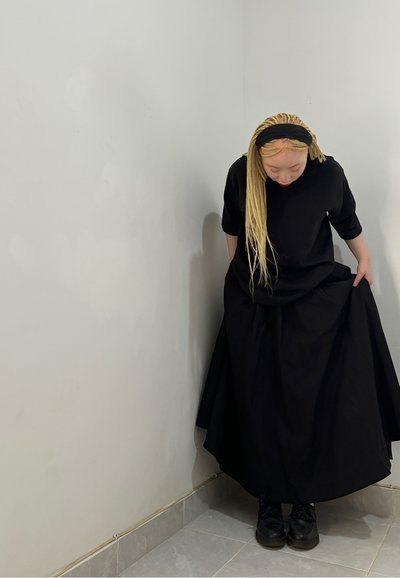 Vestido negro oversize con mangas cortas y falda fluida, combinado con zapatos negros. Textura de tela suave, de pie contra una pared gris.