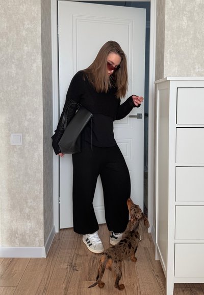 Mujer vestida de negro con gafas de sol interactuando con un perro pequeño de pelaje marrón con manchas negras en una habitación con suelo de madera y muebles blancos.