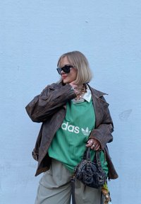 Καφέ δερμάτινο μπουφάν πάνω από ένα πράσινο φανελάκι Adidas με λευκές ρίγες και λογότυπο. Μπεζ παντελόνι με ψηλή μέση και μια μικρή σκούρα τσάντα. Μαύρα γυαλιά ηλίου.