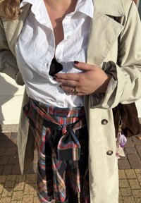 Bežinė tranšėja virš baltos marškinėlių, matomas juodas sluoksnis. Spalvinga languota sijonė, surišta juosmenyje, su juodais akcentais.