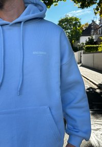 Svetlo modrá mikina s kapucňou vyrobená z jemného materiálu, má prednú kapsu a šnúrky. Malé biele logo na hrudi, voľný strih.