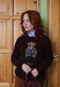 Νεαρή γυναίκα με σγουρά κόκκινα μαλλιά φοράει ένα σκούρο πουλόβερ με σχέδιο αρκούδας σε κοστούμι, κρατώντας μια μπορντό τσάντα, στέκεται δίπλα σε ξύλινη πόρτα.