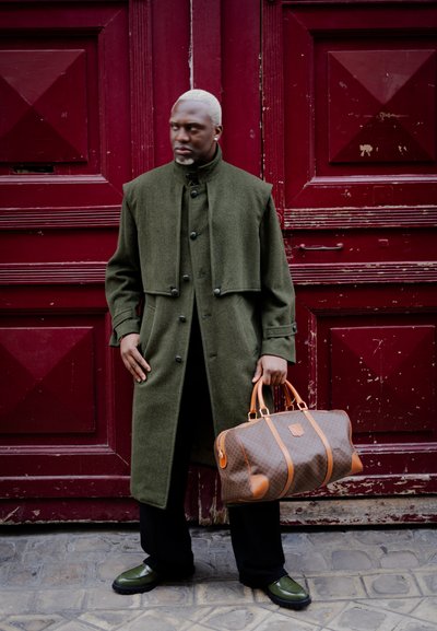 Abrigo de lana verde con cuello alto y dos botones, combinado con pantalones negros y zapatos verdes. Bolsa de viaje marrón con un diseño estampado y acentos de cuero.