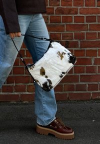 Τσάντα κύκλου σε λευκό και καφέ χρώμα με υφή μαλακής γούνας, μαύρες λεπτομέρειες και αποσπώμενο λουρί, κρατημένη από ένα άτομο που φοράει ανοιχτό μπλε τζιν.