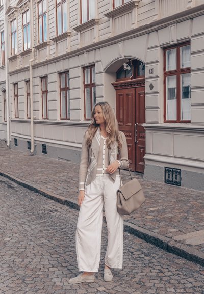 Mujer con cabello largo está de pie en una calle empedrada cerca de un edificio beige con puerta roja, lleva un cárdigan claro, pantalones blancos, mocasines y sostiene un bolso.