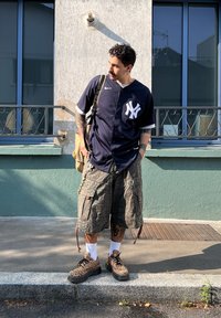 Marynarska koszulka New York Yankees, szorty w kamuflażu, i buty w panterkę. W zestawie z plecakiem, tatuażami i krótkimi, kręconymi włosami.