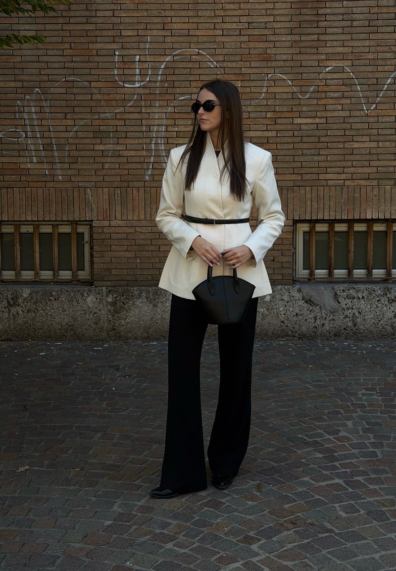 Blazer branco com cinto, calças pretas com folho e uma pequena mala preta. Óculos de sol oversized completam o visual junto a uma parede de tijolos.