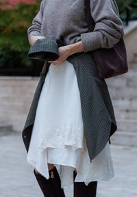 Model drži zeleno karirano srajco nad belo obleko z čipkastim robom. Rjavkaste škornje do kolen dopolnjujejo obleko. Vidna je mehka tekstura.