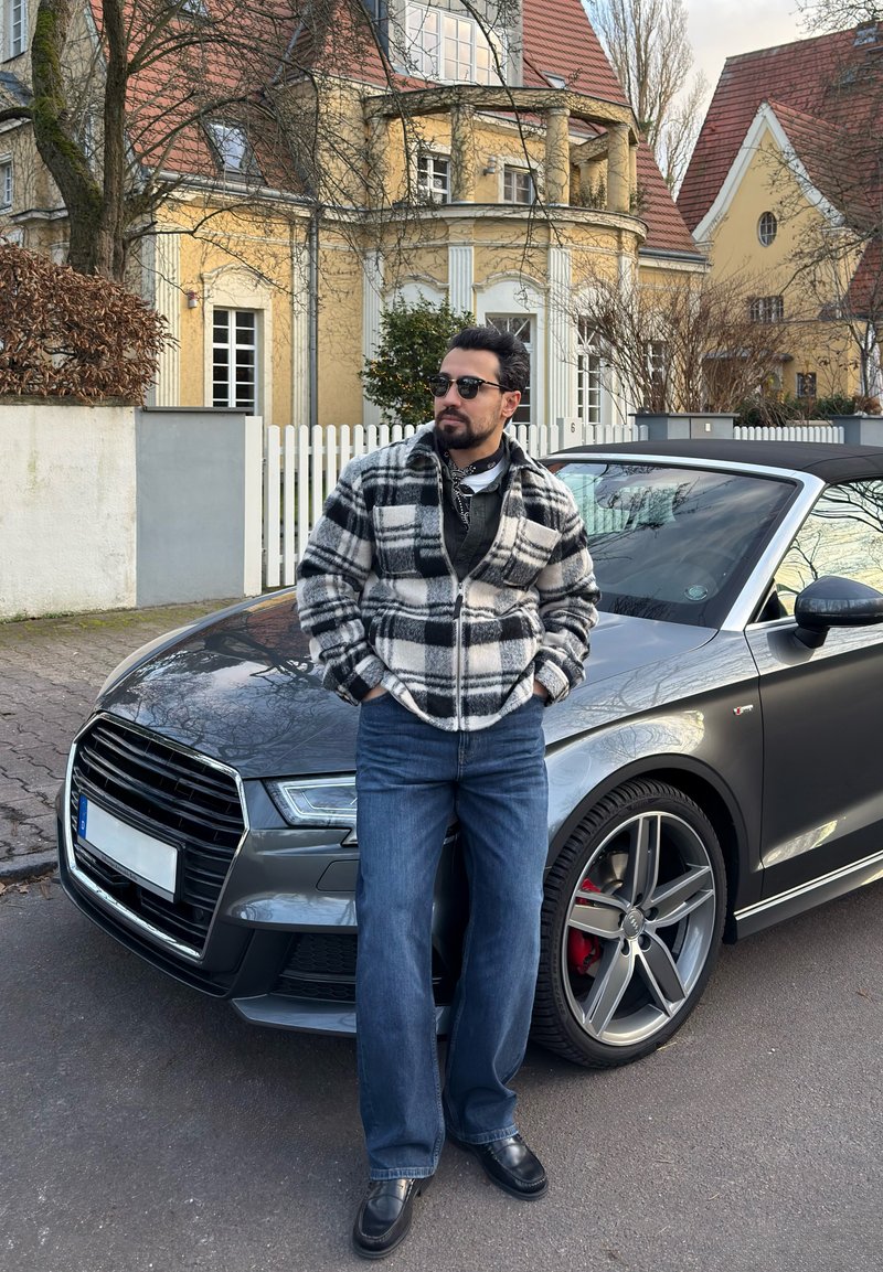 Homme portant des lunettes de soleil et une veste à carreaux, adossé à une voiture décapotable grise dans une rue avec des maisons jaunes et des clôtures blanches.