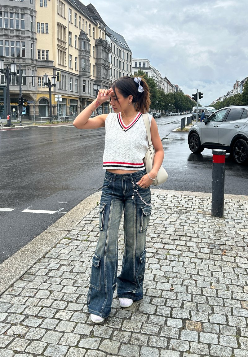 Λευκή πλεκτή κοντή μπλούζα με κόκκινες λεπτομέρειες, συνδυασμένη με φαρδιά, σκισμένα τζιν παντελόνια με τσέπες cargo και ασημένια αλυσίδα.