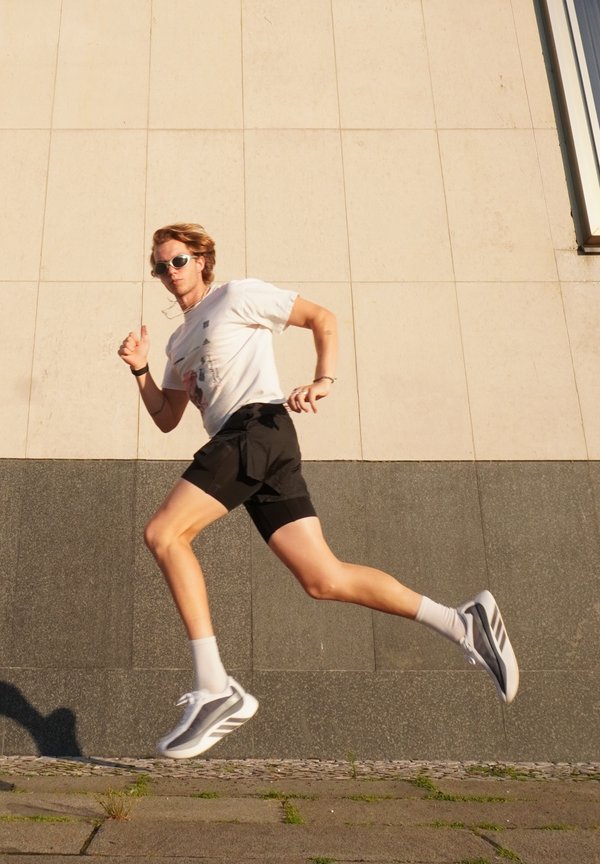 Young man wearing sunglasses and athletic clothes is mid-air running against an urban wall background in daylight.