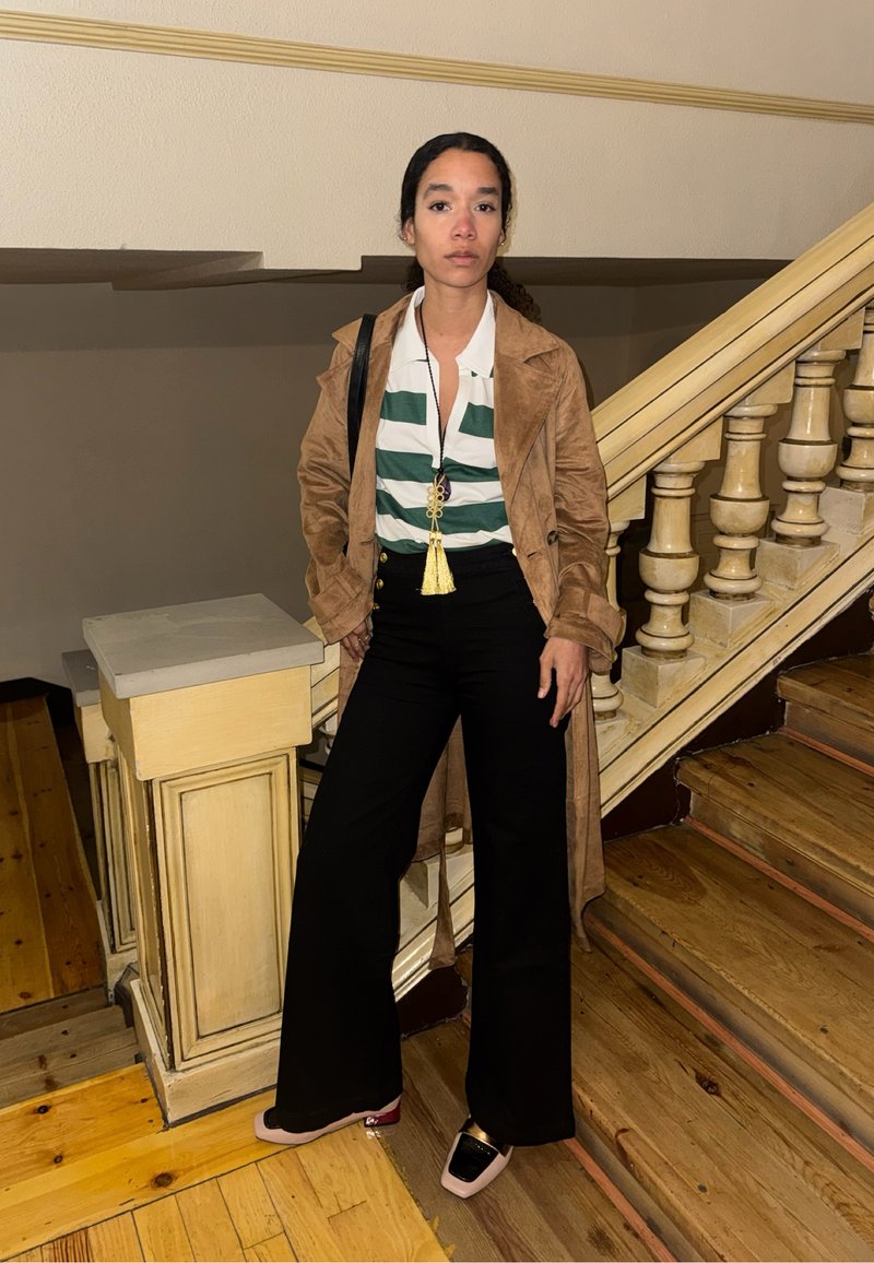 Femme aux cheveux bouclés portant un manteau marron, une chemise à rayures vertes et blanches, un pantalon noir et des mocassins, debout près d'un escalier en bois à l'intérieur.