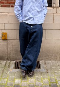 Vaaleansininen nappipaita yhdistettynä väljiin tummansinisiin farkkuihin. Yksityiskohtaiset saumat ja rento siluetti. Ruskeat nahkakengät.