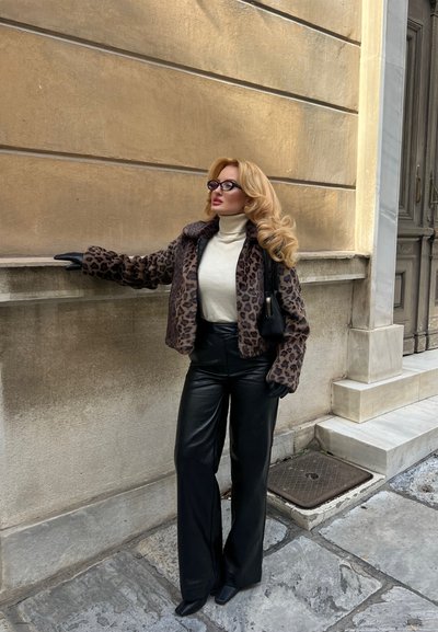 Mujer con cabello largo y rubio, que lleva un abrigo de estampado de leopardo, un suéter de cuello alto blanco, pantalones de cuero negros, gafas y guantes negros, de pie junto a una pared beige al aire libre.
