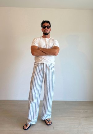 Camiseta blanca, pantalones de pierna ancha a rayas azul claro y blanca, y gafas de sol negras. De pie sobre un suelo de madera clara contra una pared blanca.