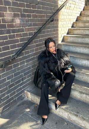 Chaqueta de piel sintética negra con mangas largas, combinada con pantalones de campana negros y zapatos negros de punta, sentado en escalones de piedra contra una pared de ladrillo.