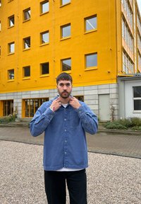 Ανοιχτό μπλε πουκάμισο με τσέπη στο στήθος και μακριά μανίκια, φορεμένο πάνω από λευκό τοπ, συνδυασμένο με μαύρο παντελόνι. Κίτρινο κτήριο στο φόντο.