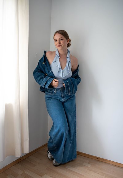 Chaqueta de mezclilla sobre un bodysuit rizado de color azul claro, combinado con jeans de pierna ancha de color azul. Zapatillas blancas sobre un suelo de madera, junto a una cortina blanca.