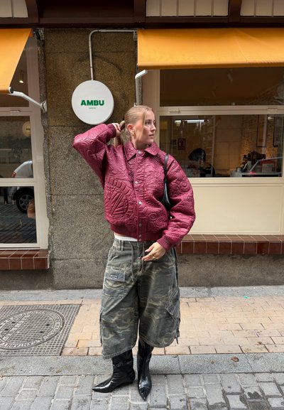 Mujer con chaqueta acolchada burdeos y pantalones anchos de camuflaje, parada frente a una cafetería con toldos amarillos.