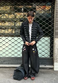 Veste de style militaire noire avec des boutons argentés, associée à un pantalon noir ample orné d'épinglettes multicolores. Chaussures marron et sac sombre.