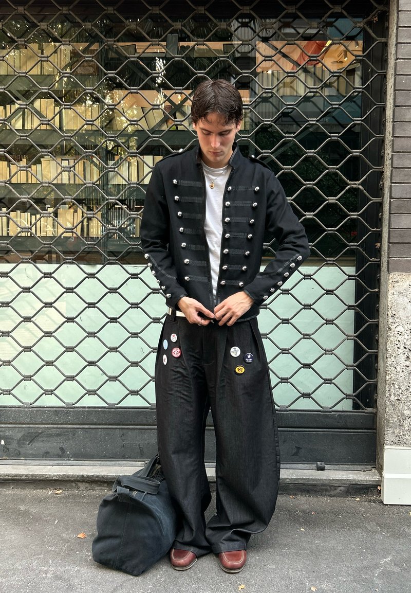 Veste de style militaire noire avec des boutons argentés, associée à un pantalon noir ample orné d'épinglettes multicolores. Chaussures marron et sac sombre.