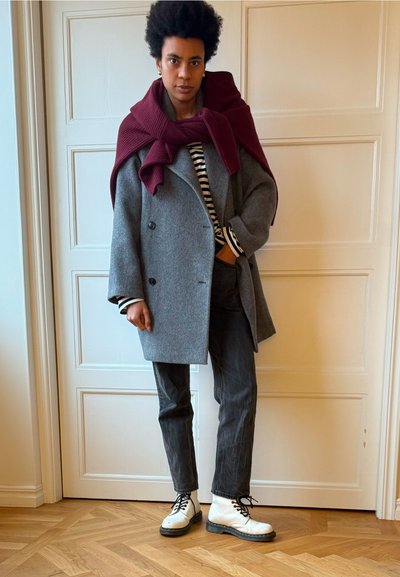 Abrigo de lana gris con doble botonadura, bufanda de punto marrón, camisa a rayas en blanco y negro, pantalones grises y botas blancas con cordones y suelas negras.