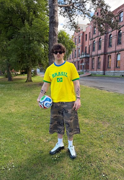 Joven con camiseta amarilla de Brasil y pantalones cortos de camuflaje sostiene un balón de fútbol de pie sobre una zona de césped cerca de un edificio de ladrillo rojo.