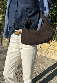 Καφέ πλεχτή τσάντα με καμπύλο σχήμα, κρατημένη από ένα άτομο που φοράει μπλε πουκάμισο με κουμπιά και λευκό παντελόνι, σε φόντο από πέτρες.