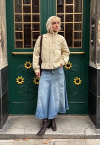 Gepolsterte beige Jacke mit Kapuze, kombiniert mit einem langen blauen Jeansrock und braunen Absatzstiefeln. Grüne Tür mit gelben Sonnenblumenakzenten.