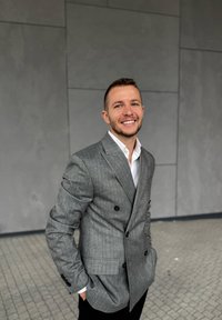 Szary garnitur w prążki podwójnie zapinany, z czarnymi guzikami, noszony na białej koszuli, o nowoczesnym kroju i gładkiej fakturze. Czarne spodnie.