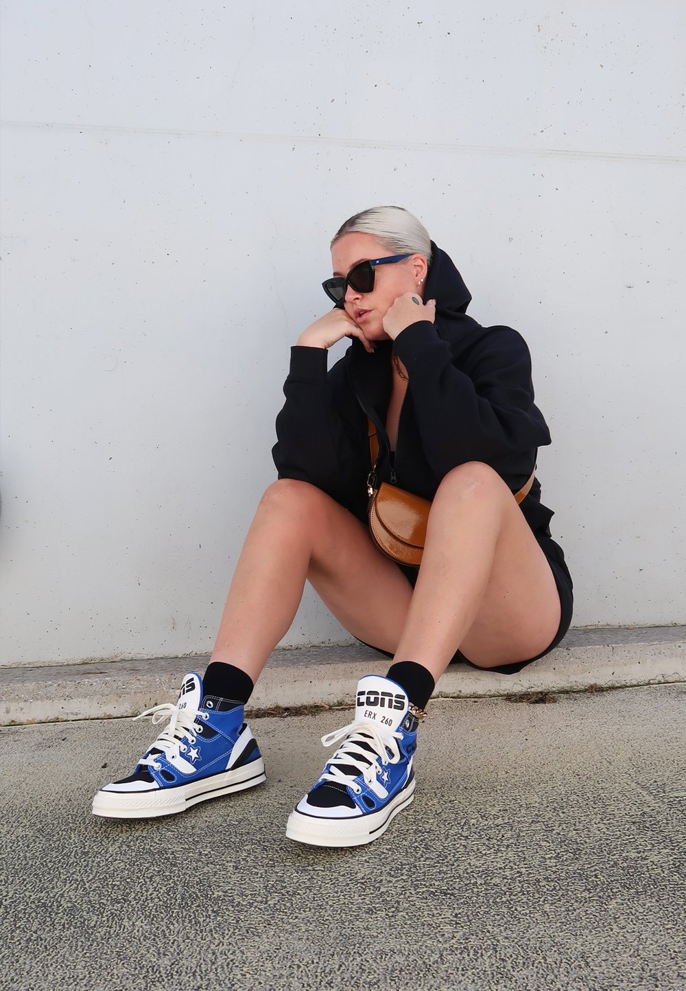 Zwarte rits hoodie, zwarte shorts, blauwe high-top sneakers met witte accenten, zwarte en blauwe sokken, en een kleine bruine schoudertas.