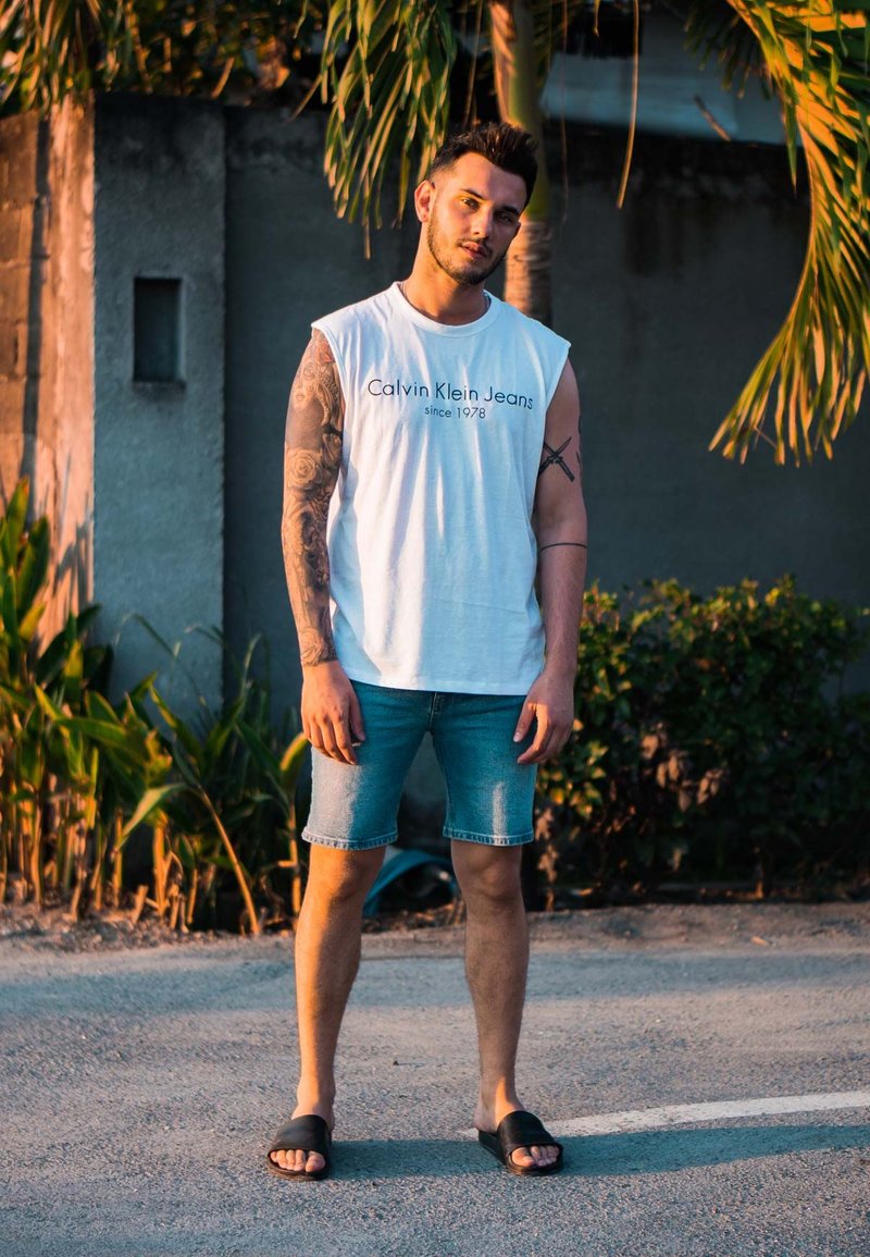 Weißes ärmelloses Hemd mit "Calvin Klein Jeans"-Branding, Denim-Shorts und schwarzen Slidesandalen; steht auf einem Betonweg mit Grünpflanzen.