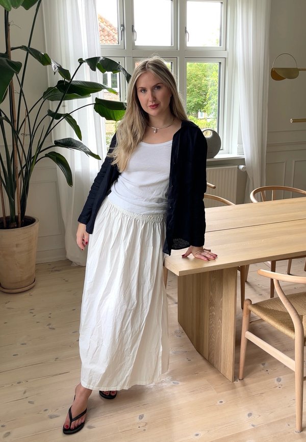 Witte, vloeiende rok met een geraffineerde taille, gecombineerd met een aansluitend wit topje en een zwarte overhemd met knopen. De scène bevat een houten tafel en een plant.