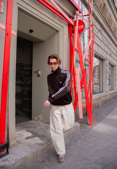 Persona joven con gafas de sol, chaqueta deportiva negra y pantalones blancos caminando por la acera urbana junto a arte metálico rojo y la entrada de un edificio.