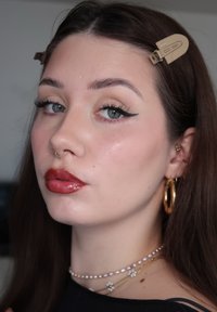 Jeune femme aux cheveux foncés, avec un trait d'eyeliner façon aile, un rouge à lèvres rouge, un anneau de septum en or, des boucles d'oreilles créoles en or, et des colliers superposés, tournée légèrement vers la gauche.
