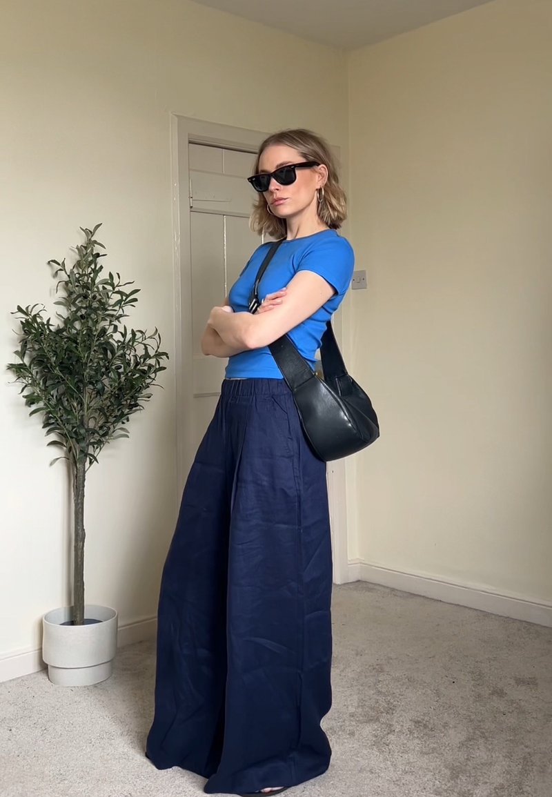 Frau mit Sonnenbrille, blauem Kurzarmhemd, weiten marineblauen Hosen und schwarzer Umhängetasche steht mit verschränkten Armen in einem Zimmer neben einer Topfpflanze.