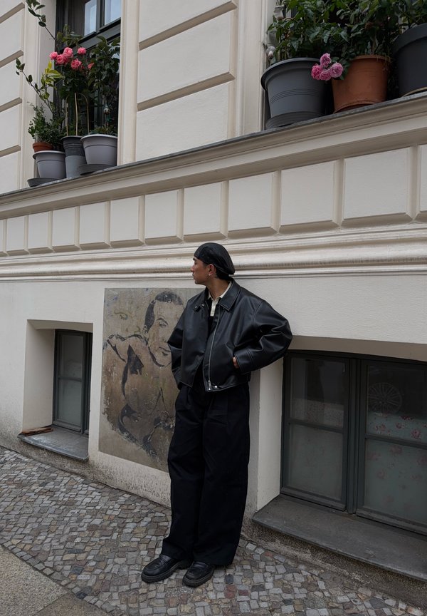 Person in schwarzer Lederjacke und Hose steht auf einer Kopfsteinpflasterstraße und lehnt an einer Wand mit einem gemalten Porträt und Fenstern darüber.