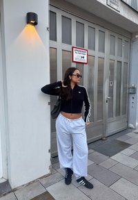 Fekete zipzáros crop top fehér csíkokkal, szürke bő szabású melegítőnadrág és fekete cipő fehér kiegészítőkkel. Egy üvegajtó mellett áll.